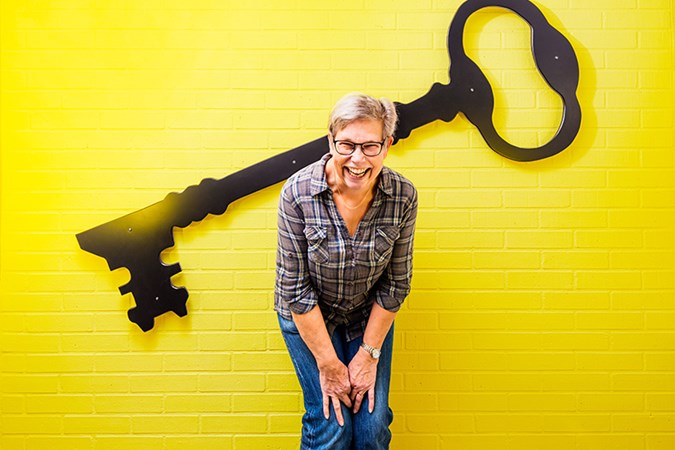 Administrativ assistent Anki Gävert framför en stor svart nyckel som hänger på en gul vägg.