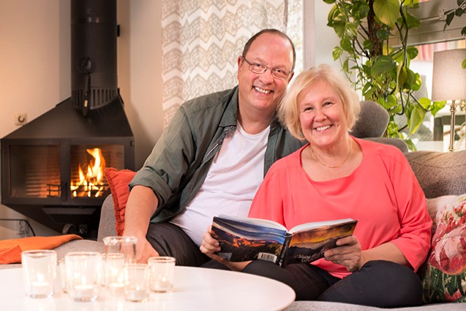 Jörgen och Katarina sitter i soffan i vardagsrummet. Braskaminen brinner i bakgrunden och det är varma färger i bilden.