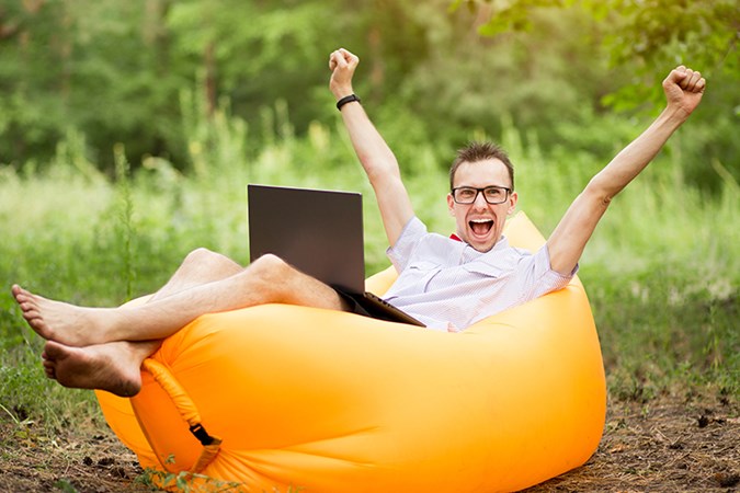 Lycklig man som sitter utomhus på en uppblåst säck med laptop i knät.